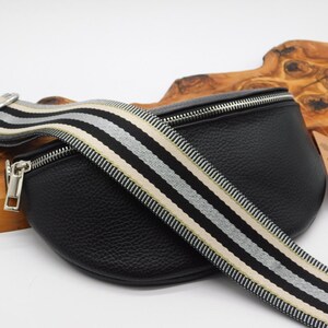 May include: Black leather bum bag with a striped strap in black, white, and beige. The bag has a silver zip and hardware. The strap is adjustable. The bag is on a white surface.