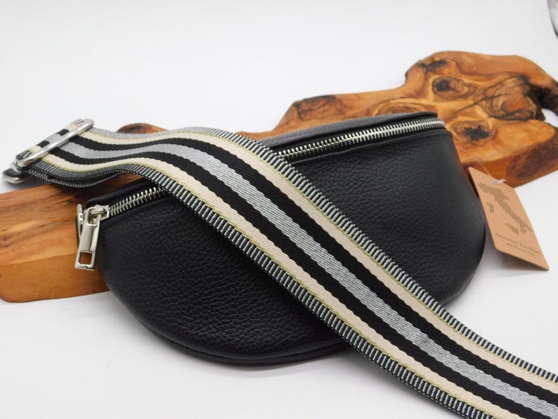 May include: Black leather bum bag with a striped strap in black, white, and beige. The bag has a silver zip and hardware. The strap is adjustable. The bag is on a white surface.