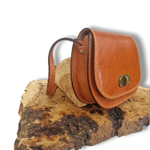 May include: A small, tan leather bag with a curved flap and a brass-coloured clasp. The bag has a stitched handle and is displayed on a piece of textured wood. The bag is a saddle style.
