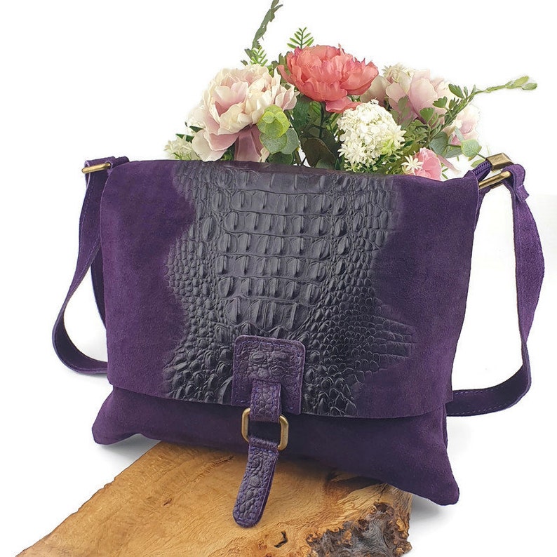 May include: A purple suede crossbody bag with a flap featuring a dark, textured, crocodile-patterned panel. The bag has a long strap, a buckle closure, and gold-colored hardware. The bag is approximately 10 inches wide.