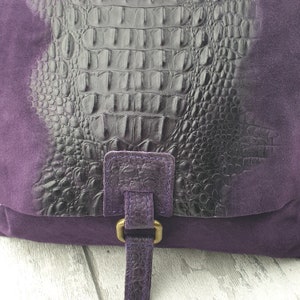 May include: A purple handbag with a flap featuring a black, textured crocodile-print design. The bag has a suede-like texture on the sides and a strap with a gold-colored buckle. The bag is designed for carrying items.