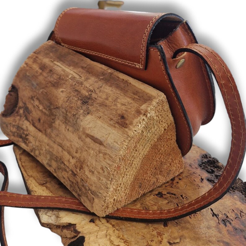 May include: A small, brown leather crossbody bag with a long strap. The bag has a rectangular shape with a flap closure and a brass-coloured clasp. The bag is resting on a piece of wood.