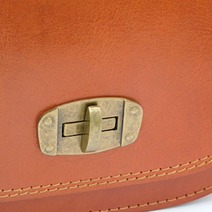 May include: Close-up of a tan leather bag featuring a brass-toned clasp. The bag's edges are defined by visible stitching, and the leather has a textured appearance. The clasp is rectangular with a central lever.