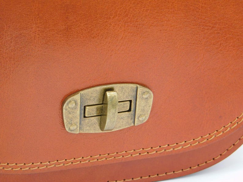May include: Close-up of a tan leather bag featuring a brass-toned clasp. The bag's edges are defined by visible stitching, and the leather has a textured appearance. The clasp is rectangular with a central lever.