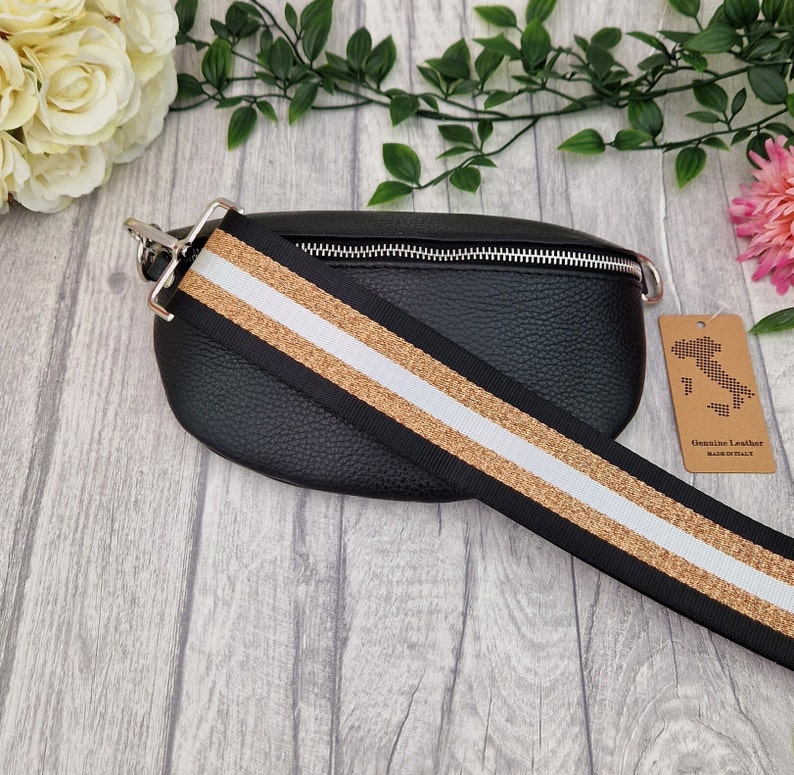 May include: Black leather bum bag with a silver zip and a wide strap with black, gold, and white stripes. The bag has a textured finish and a tag reading "Genuine Leather Made in Italy."