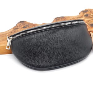 May include: Black leather bum bag with a silver zip. The bag has a textured surface and a curved shape. A small tag is attached to the side. The bag is displayed on a wooden background.