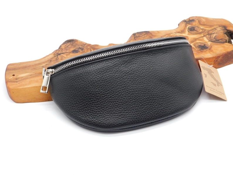May include: Black leather bum bag with a silver zip. The bag has a textured surface and a curved shape. A small tag is attached to the side. The bag is displayed on a wooden background.