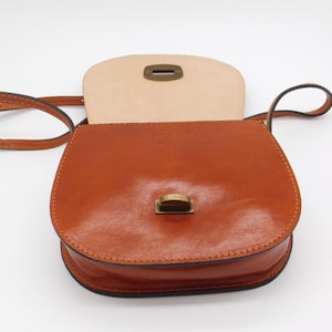 May include: A tan leather crossbody bag with a flap closure and a long strap. The bag has a rounded shape and a brass-coloured clasp. The interior is a lighter shade of tan leather. The bag is designed for carrying essentials.
