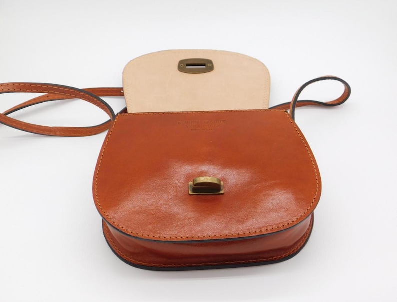 May include: A tan leather crossbody bag with a flap closure and a long strap. The bag has a rounded shape and a brass-coloured clasp. The interior is a lighter shade of tan leather. The bag is designed for carrying essentials.