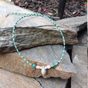 May include: A turquoise, white, and pink beaded necklace with a turquoise pendant. The necklace is on a pile of rocks.