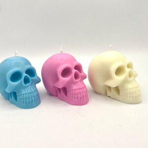 May include: Three skull-shaped candles in different colors: light blue, pink, and white. The candles are made of wax and have a realistic skull design.