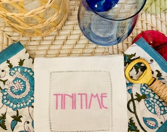 May include: White linen cocktail napkin with pink embroidery that reads "TINI TIME". The napkin is on a woven wicker surface with a blue glass, a gold bottle opener, and a floral patterned napkin.