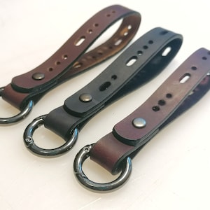 May include: Three leather keyrings with metal rings. The keyrings are brown, black, and burgundy. Each keyring has a stitched design and a metal button.