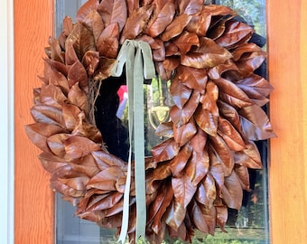 Peut inclure: Une couronne circulaire composée de feuilles de magnolia séchées brun foncé, ornée d'un ruban de velours vert sauge clair noué en boucle au centre supérieur. La couronne est fixée sur une porte vitrée où le numéro 201 est visible.