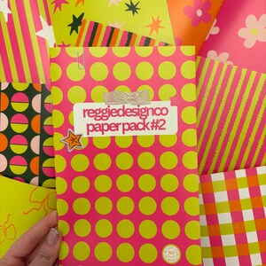 May include: A collection of colorful patterned papers. The central paper pack, labeled "reggiedesignco paper pack #2", features a pink background with large yellow polka dots. Other papers display stripes, stars, and floral designs in vibrant pink, orange, and green.