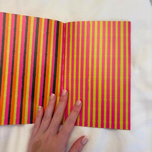 May include: Open book with colorful vertical stripes in neon pink, yellow, orange, and black. The book is open, revealing the patterned pages. A hand rests on the book.