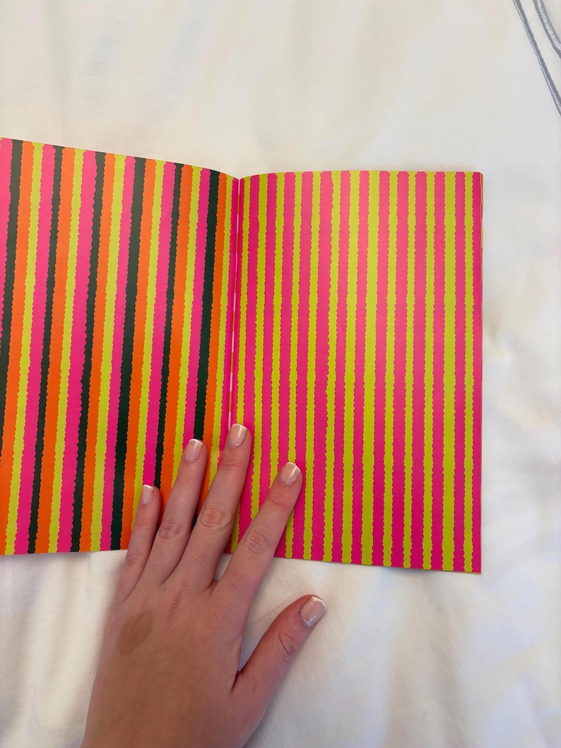 May include: Open book with colorful vertical stripes in neon pink, yellow, orange, and black. The book is open, revealing the patterned pages. A hand rests on the book.