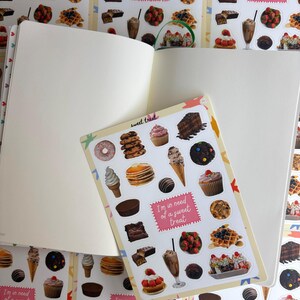 May include: An open notebook with blank pages and a sticker sheet displaying an assortment of desserts, including donuts, cupcakes, and ice cream. The sticker sheet has the text "sweet treat" and "I'm in need of a sweet treat."