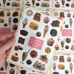 May include: A sheet of colourful stickers featuring various desserts such as doughnuts, cupcakes, ice cream cones, and chocolate candies. The stickers have the text "I'm in need of a sweet treat" and "sweet treat".