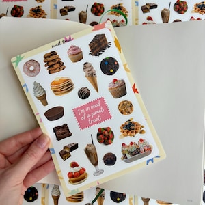 May include: A sticker sheet with illustrations of desserts like cupcakes, ice cream, and waffles. The text "I'm in need of a sweet treat" is on a pink label. The sheet is held over an open notebook, with a repeating pattern of the stickers.