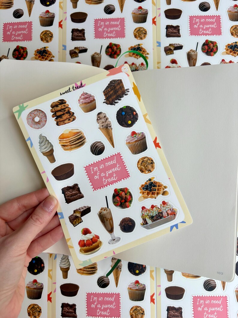 May include: A sticker sheet with illustrations of desserts like cupcakes, ice cream, and waffles. The text "I'm in need of a sweet treat" is on a pink label. The sheet is held over an open notebook, with a repeating pattern of the stickers.