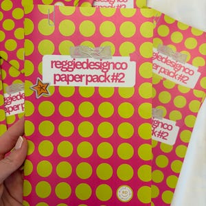 May include: A stack of pink paper packs with a repeating pattern of large yellow circles. Each pack is labeled with the text "reggiedesignco paper pack #2" and secured with a paper clip and tape. A star sticker is visible.