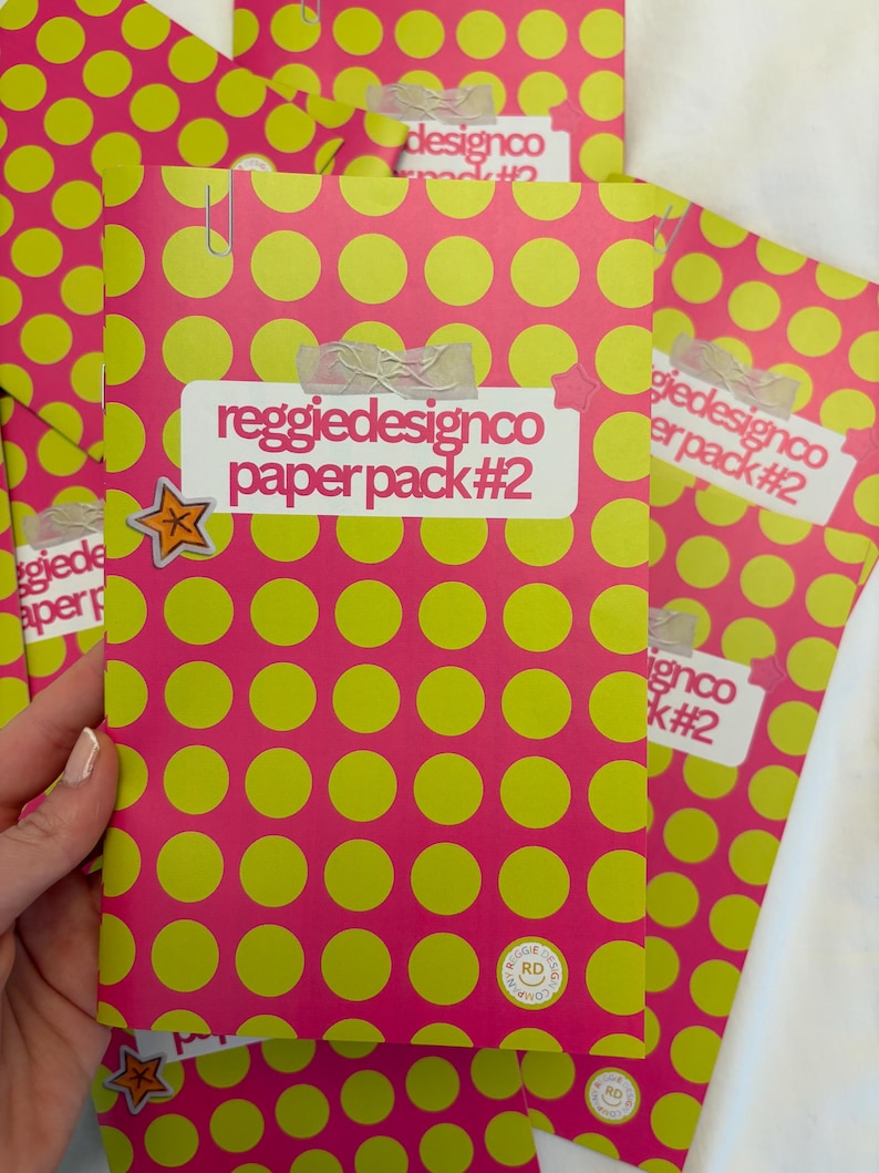 May include: A stack of pink paper packs with a repeating pattern of large yellow circles. Each pack is labeled with the text "reggiedesignco paper pack #2" and secured with a paper clip and tape. A star sticker is visible.