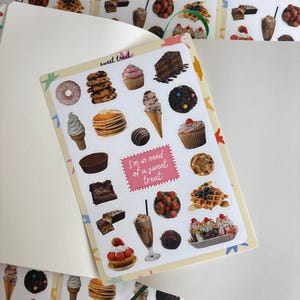May include: A sticker sheet with illustrations of sweet treats such as cupcakes, ice cream, and waffles. The stickers have a light pink border and the text "I'm in need of a sweet treat". The stickers are displayed on a white background.