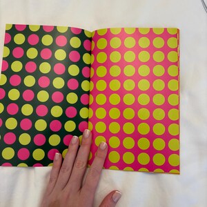 May include: Open book with pages featuring colorful polka dot patterns. One page has a dark green background with pink and yellow dots. The other page has a pink background with yellow dots. The book is open on a white surface.