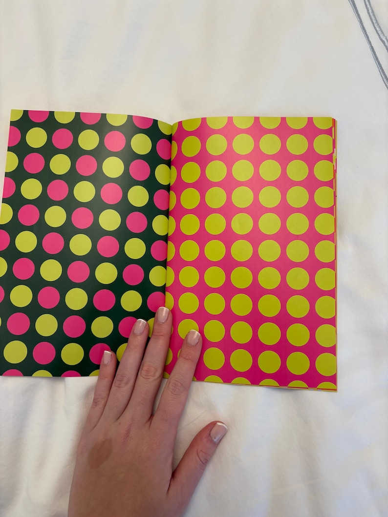 May include: Open book with pages featuring colorful polka dot patterns. One page has a dark green background with pink and yellow dots. The other page has a pink background with yellow dots. The book is open on a white surface.