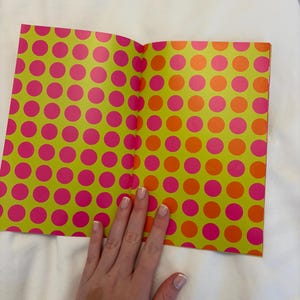 May include: Open notebook with a lime green cover featuring a pattern of large pink and orange polka dots. The notebook is open, revealing the inside pages. A hand rests on the pages.