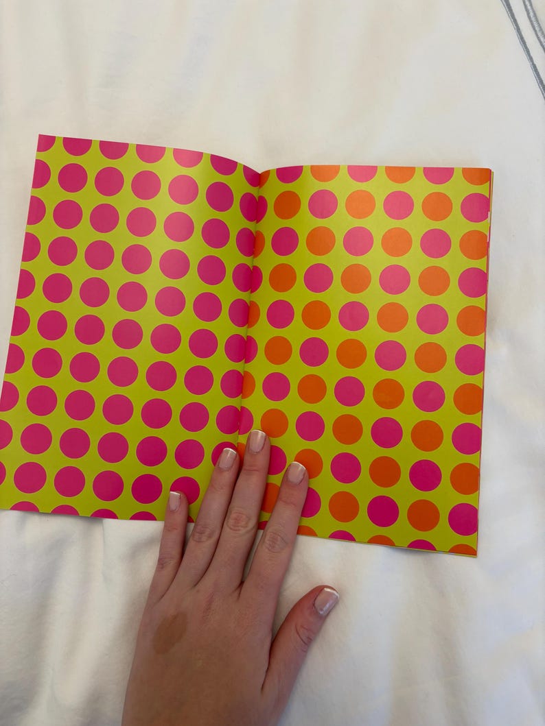 May include: Open notebook with a lime green cover featuring a pattern of large pink and orange polka dots. The notebook is open, revealing the inside pages. A hand rests on the pages.