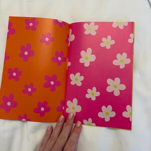 May include: Open book with orange and pink pages featuring a floral pattern. The left page is orange with pink flowers, and the right page is pink with white flowers. The book is open on a white surface.