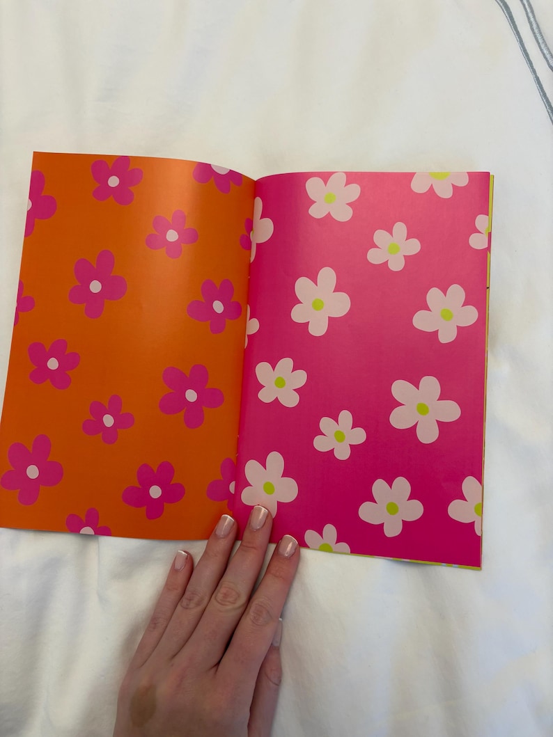 May include: Open book with orange and pink pages featuring a floral pattern. The left page is orange with pink flowers, and the right page is pink with white flowers. The book is open on a white surface.