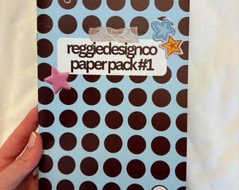 May include: A paper pack with a light blue cover featuring a pattern of brown polka dots. The text "reggiedesignco paper pack #1" is visible. Decorative star and paperclip embellishments are attached to the cover.