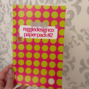 May include: A pink paper pack with a pattern of large yellow circles. The text "reggiedesignco paper pack #2" is on a white label. A star-shaped sticker and a paper clip are attached to the pack.