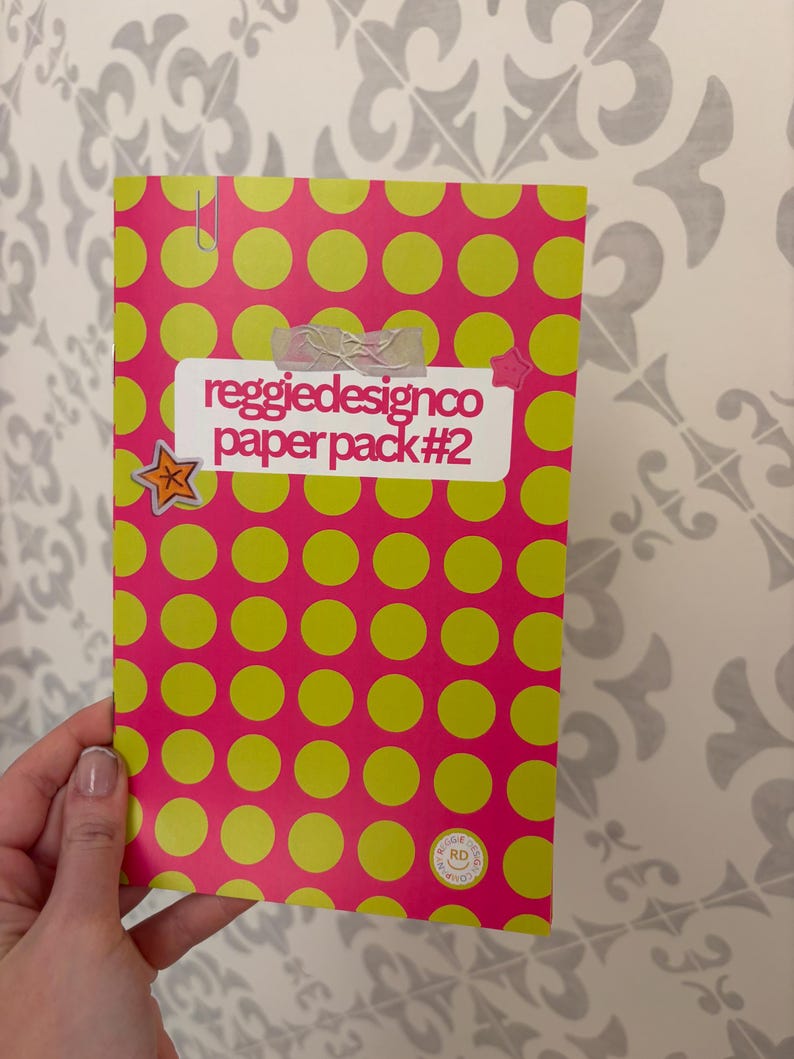 May include: A pink paper pack with a pattern of large yellow circles. The text "reggiedesignco paper pack #2" is on a white label. A star-shaped sticker and a paper clip are attached to the pack.