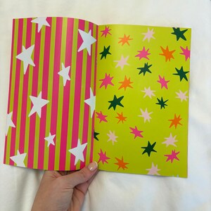 May include: Open book with colorful star patterns. One page features white stars on pink and green striped background. The other page has stars in pink, orange, and green on a light green background. The book is held by a hand.