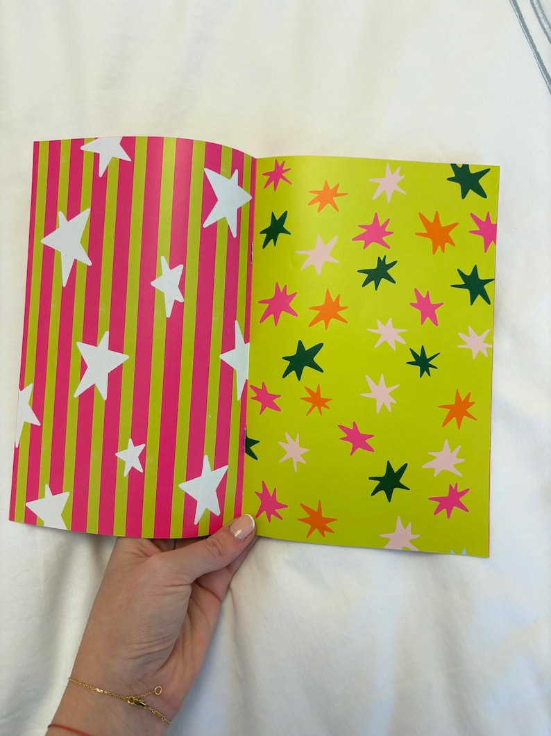 May include: Open book with colorful star patterns. One page features white stars on pink and green striped background. The other page has stars in pink, orange, and green on a light green background. The book is held by a hand.