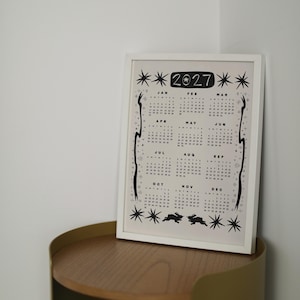 May include: A framed 2027 calendar print featuring black ink illustrations of stars, celestial motifs, and two rabbits at the bottom. The calendar displays all twelve months from January to December in a grid layout with a white border and frame.