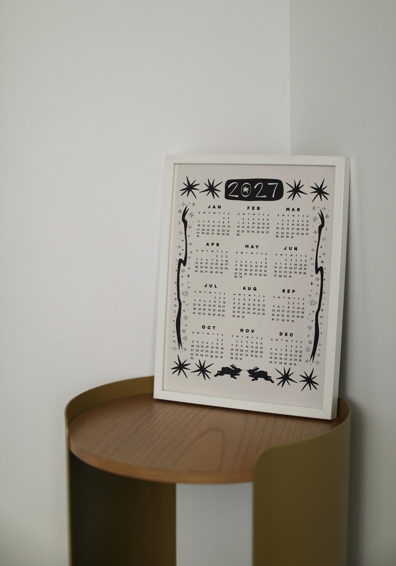May include: A framed 2027 calendar print featuring black ink illustrations of stars, celestial motifs, and two rabbits at the bottom. The calendar displays all twelve months from January to December in a grid layout with a white border and frame.