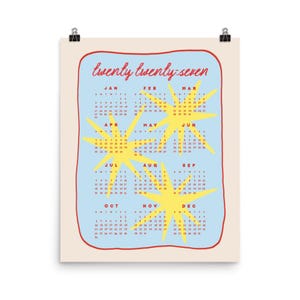 May include: A 2027 wall calendar on a light cream background featuring a light blue rectangular area with a red hand-drawn border. The text twenty twenty-seven is written in red script above twelve months arranged in grids with yellow star graphics.