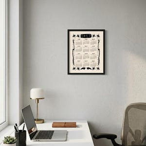 May include: A framed 2027 wall calendar with a cream background and black ink illustrations. The design features the year 2027 at the top, twelve monthly grids, star motifs, and two rabbit silhouettes at the bottom, all enclosed in a black frame.