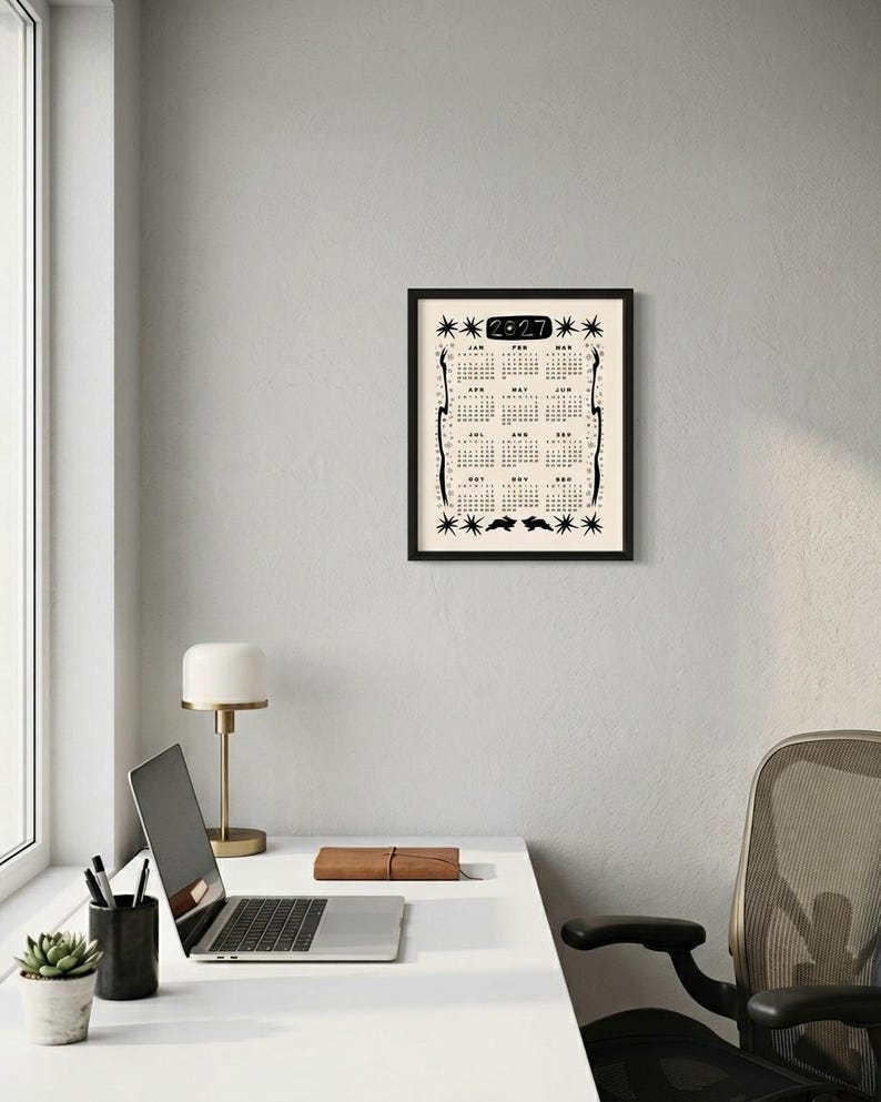 May include: A framed 2027 wall calendar with a cream background and black ink illustrations. The design features the year 2027 at the top, twelve monthly grids, star motifs, and two rabbit silhouettes at the bottom, all enclosed in a black frame.