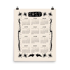 May include: A 2027 wall calendar printed on a cream background with black ink. The design features twelve monthly grids, star motifs, and two rabbit silhouettes at the bottom. The year 2027 is displayed in a black rounded rectangular header.