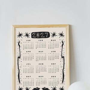 May include: A 2027 wall calendar in a light wood frame featuring a cream background with black ink illustrations of stars, celestial motifs, and two rabbits at the bottom. The calendar displays all twelve months with dates and days of the week.