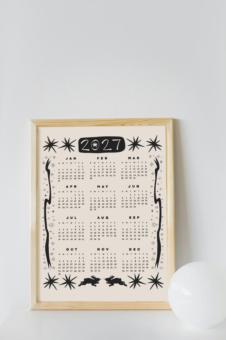 May include: A 2027 wall calendar in a light wood frame featuring a cream background with black ink illustrations of stars, celestial motifs, and two rabbits at the bottom. The calendar displays all twelve months with dates and days of the week.