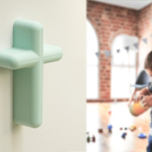 Peut inclure: Une décoration murale en forme de croix en plastique vert clair aux bords arrondis. La croix est fixée à un mur blanc.