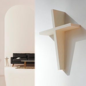 Peut inclure: Une étagère en bois blanc en forme de croix, montée sur un mur blanc. L'étagère a deux niveaux et est légèrement inclinée vers l'avant.