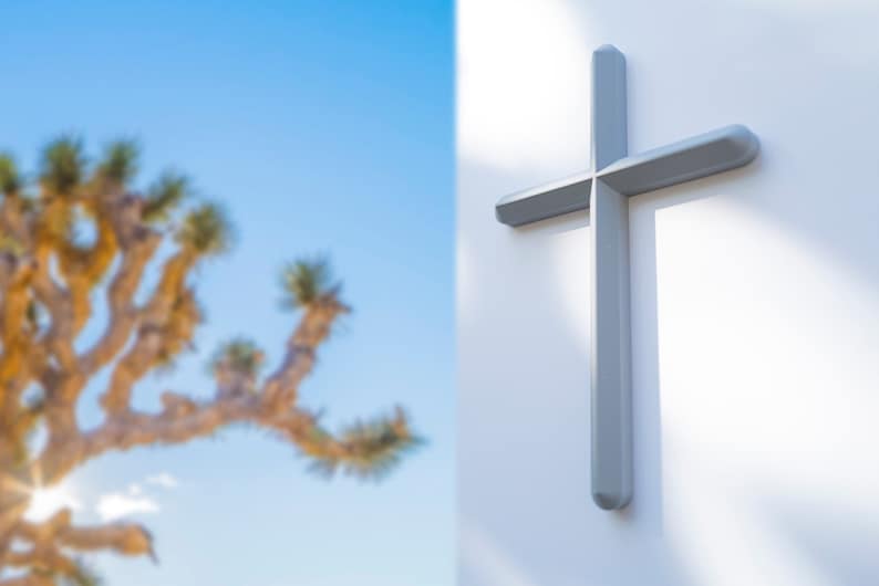 May include: A silver cross mounted on a white wall. The cross is a simple design with a straight beam and a vertical post. The background is out of focus and shows a desert landscape.